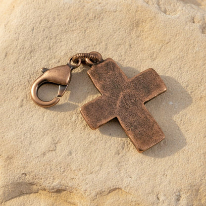 Hackberry Creek distressed copper cross charm with a lobster clasp, resting on a textured beige stone surface.