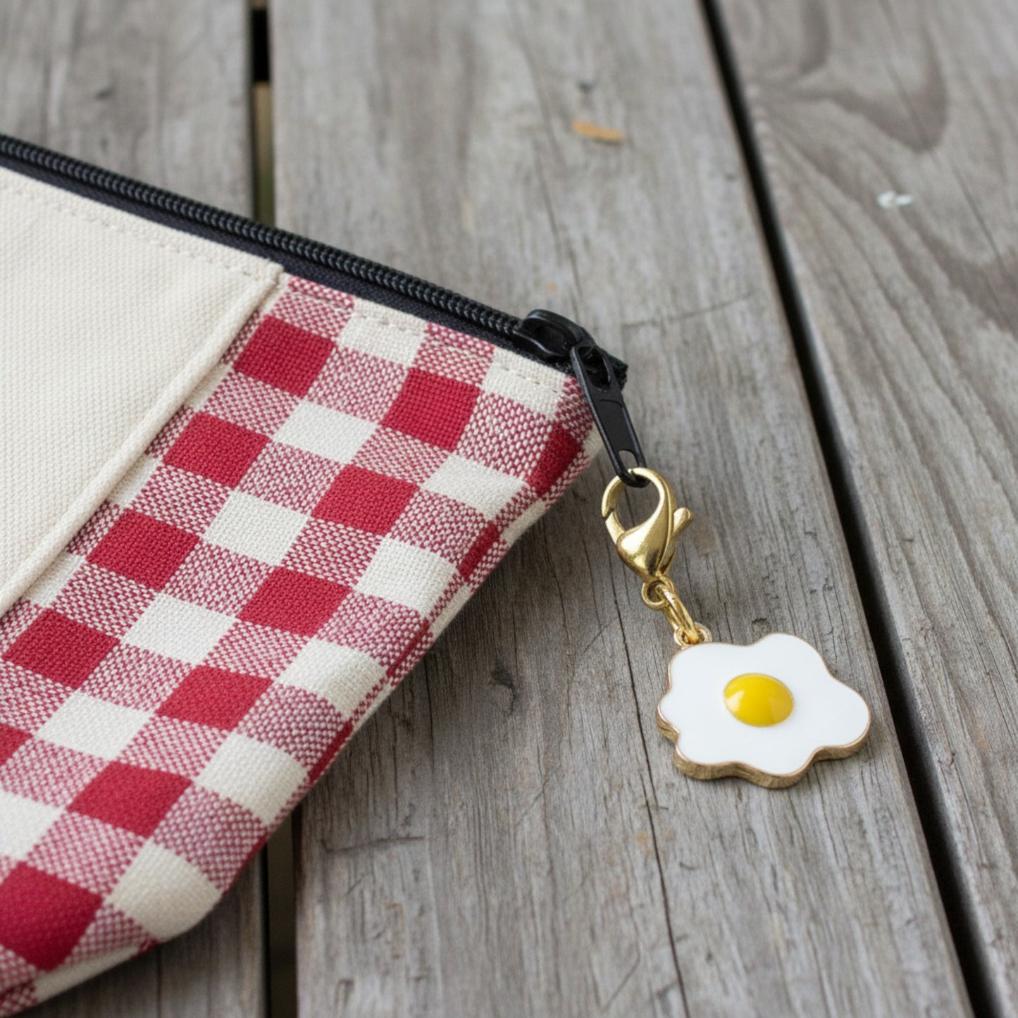 Hackberry Creek enamel fried egg charm with a gold lobster clasp attached to a red and white gingham zipper pouch.
