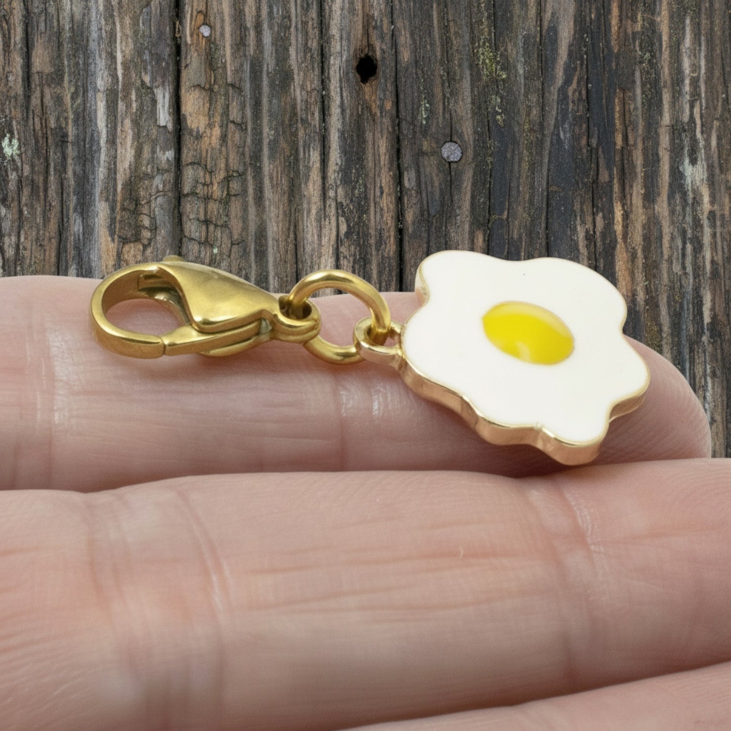 Hackberry Creek enamel fried egg charm with a yellow yolk and gold-plated lobster clasp held in a hand.