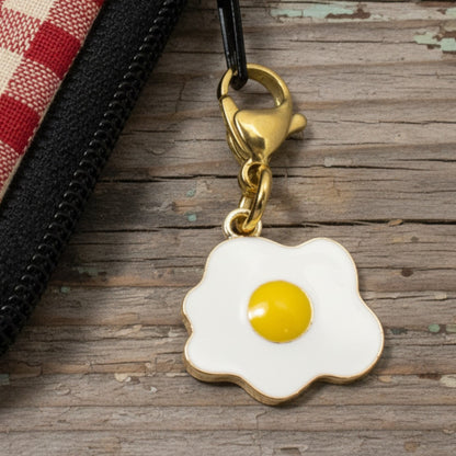 Hackberry Creek enamel fried egg charm with a gold lobster clasp attached to a red and white gingham zipper pouch.