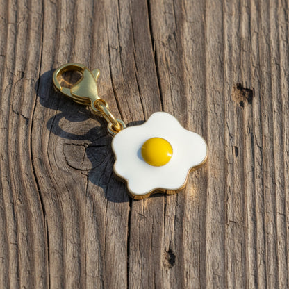 Hackberry Creek enamel fried egg charm with a yellow yolk and gold-plated lobster clasp on a rustic wood background.