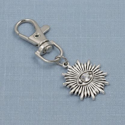 Hackberry Creek silver metal alloy evil eye sunburst bag charm with a clear rhinestone accent on a blue fabric background