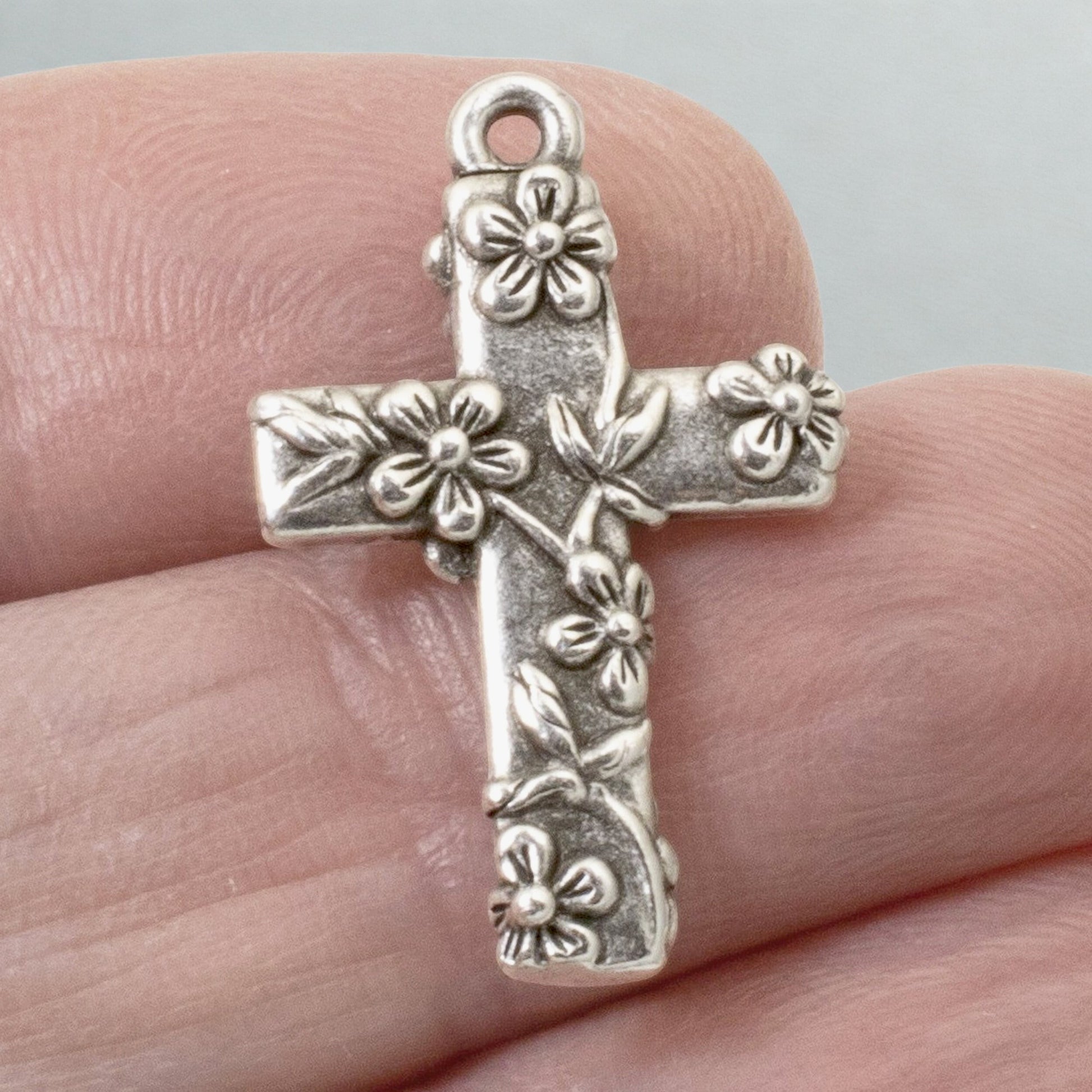 Hackberry Creek antique silver-plated pewter floral cross pendant held between fingers for scale.