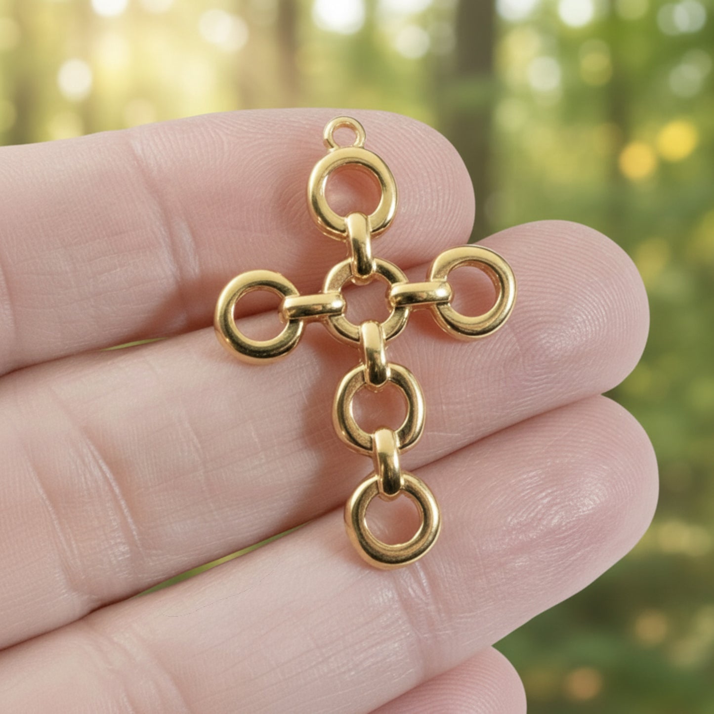 Hackberry Creek 24K gold plated open chain-link cross pendant held between fingers against a soft green background.