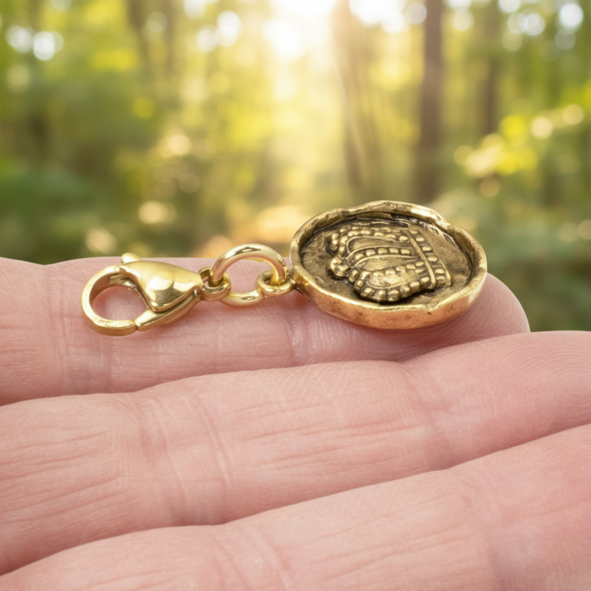 Hackberry Creek antique gold crown charm with a lobster clasp held in a hand against a blurred forest background.