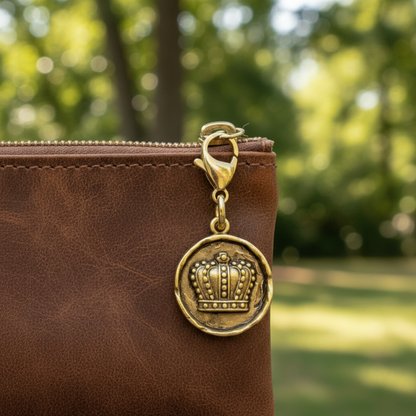 Hackberry Creek antique gold crown charm with lobster clasp attached to a brown leather bag in a garden setting