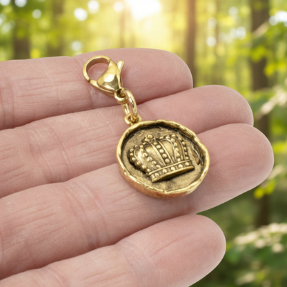 Hackberry Creek gold crown charm with an antique finish and lobster clasp held in a hand against a blurred forest.