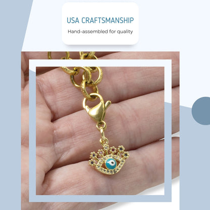 Hackberry Creek gold evil eye charm with blue enamel and colorful CZ accents on a lobster clasp held in a hand.