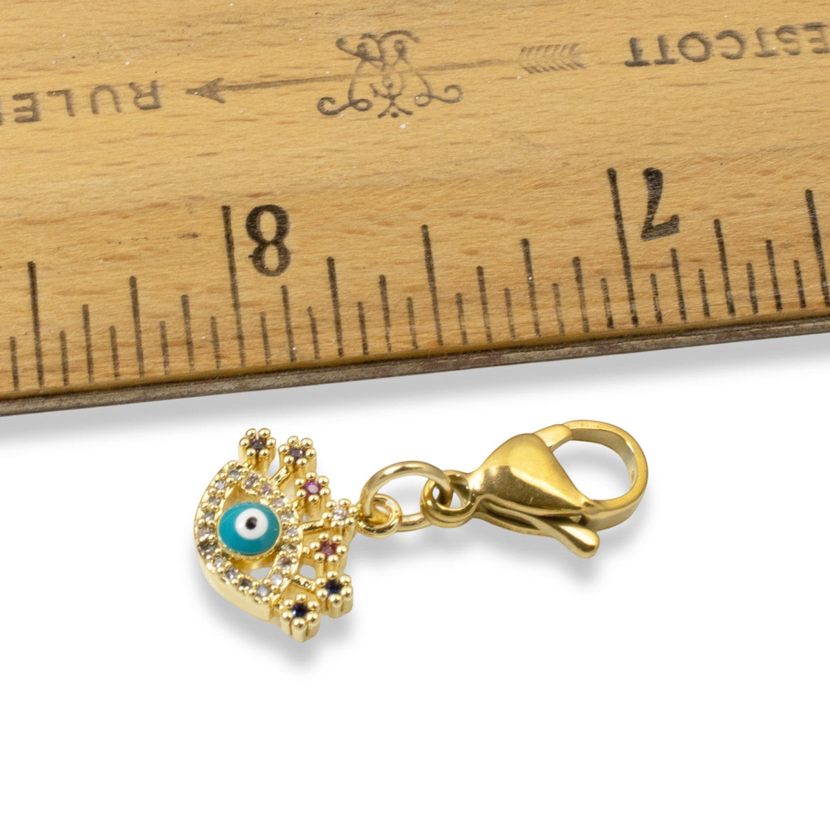 Hackberry Creek gold evil eye charm with blue enamel, cubic zirconia accents, and lobster clasp next to a ruler