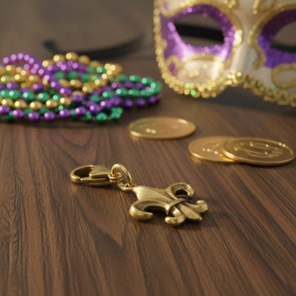 Hackberry Creek 24k gold-plated Fleur de Lis charm on a wooden surface with Mardi Gras beads and a masquerade mask.