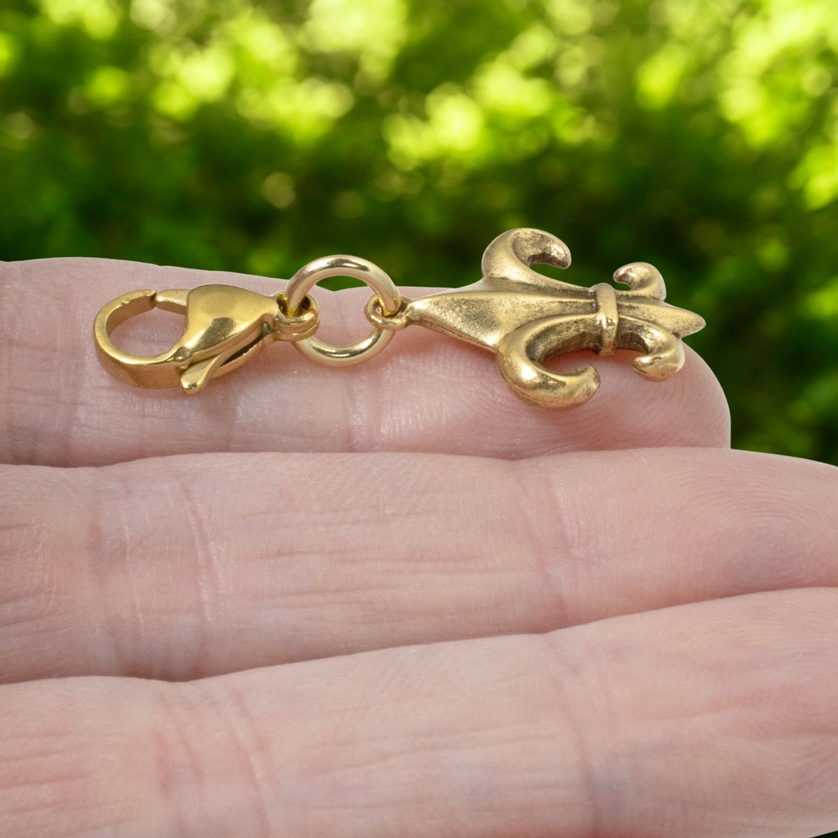Hackberry Creek 24k gold-plated Fleur de Lis charm with a secure clasp held in a hand against a blurred green background