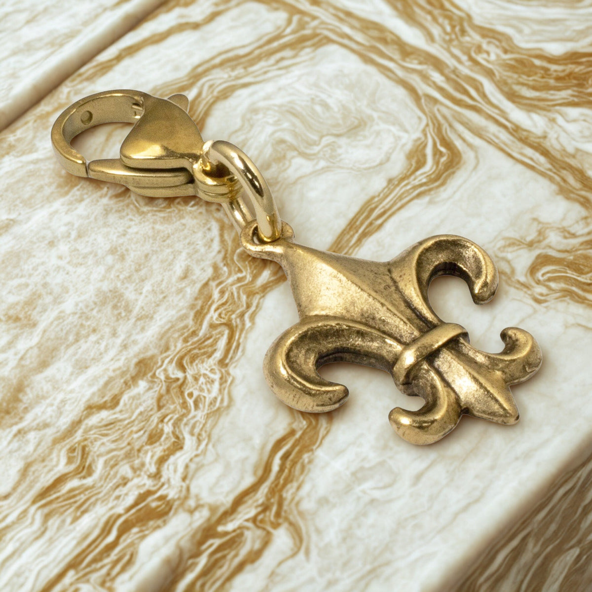 Hackberry Creek 24k gold-plated Fleur de Lis charm with a secure clasp, resting on a marbled surface.