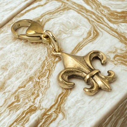 Hackberry Creek 24k gold-plated Fleur de Lis charm with a secure clasp, resting on a marbled surface.