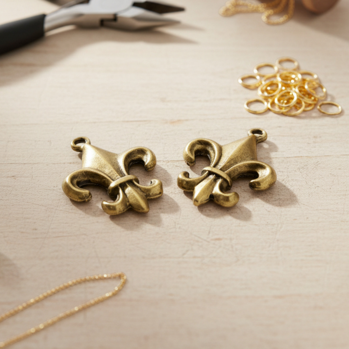 Two Hackberry Creek gold fleur de lis charms with an antique finish, displayed on a wooden jewelry making surface.