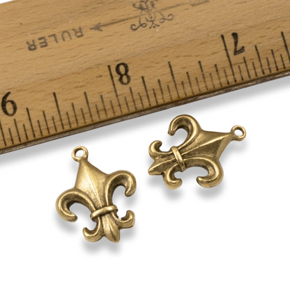 Hackberry Creek gold fleur de lis charms with antique finish next to a wooden ruler for scale