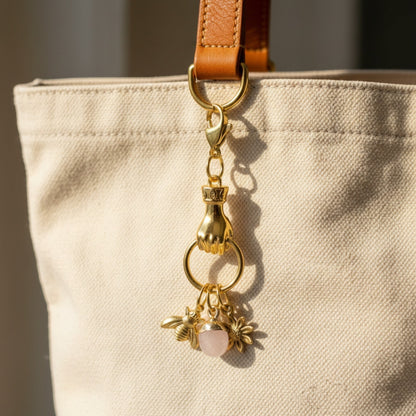 Hackberry Creek gold plated hand charm holder with bee, flower, and rose quartz charms attached to a canvas bag.