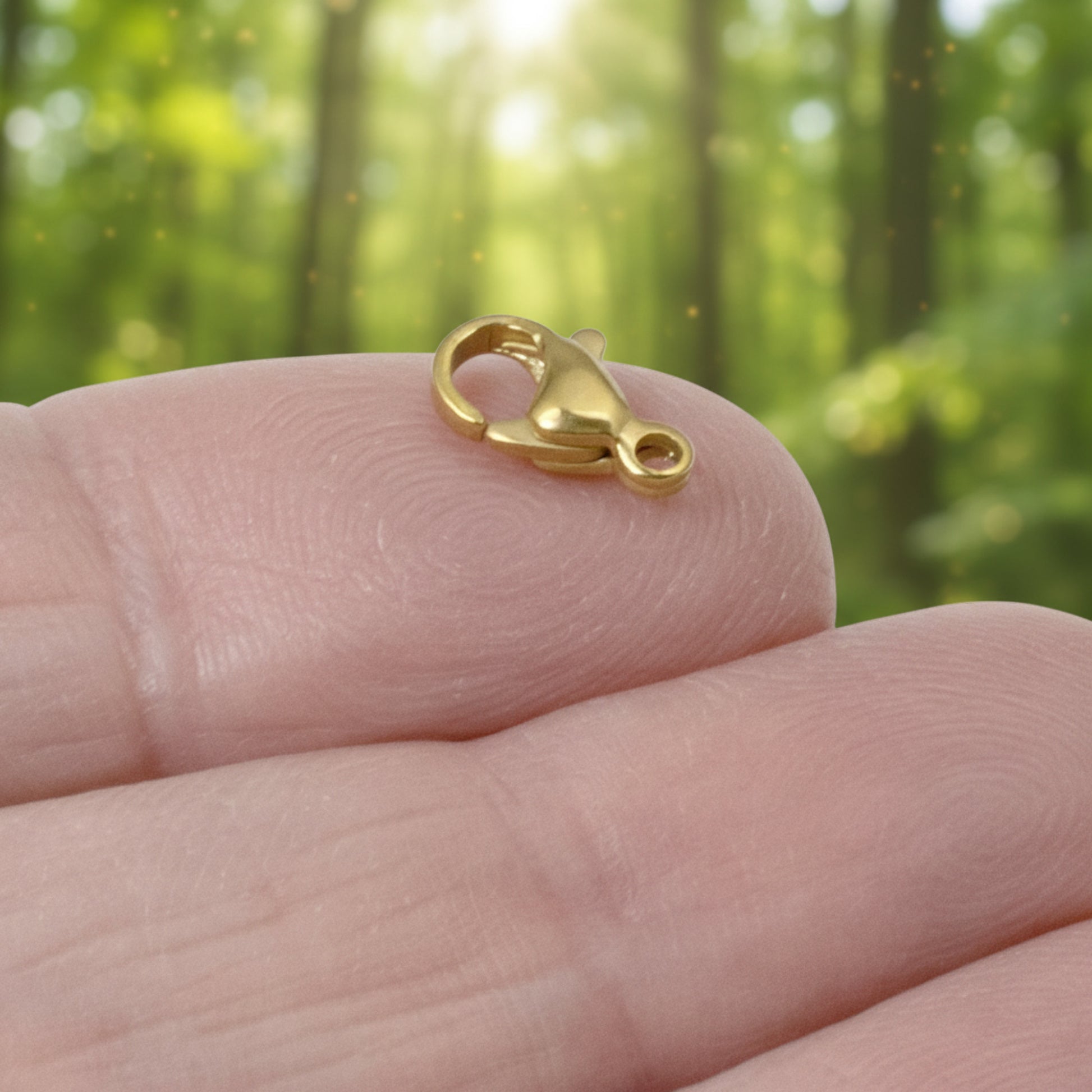 Hackberry Creek 18k gold-plated stainless steel lobster claw clasp resting on a fingertip against a forest background