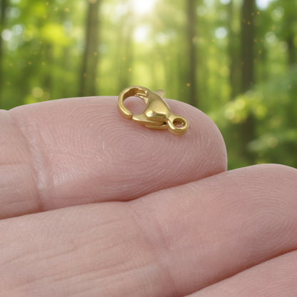 Hackberry Creek 18k gold-plated stainless steel lobster claw clasp resting on a fingertip against a forest background