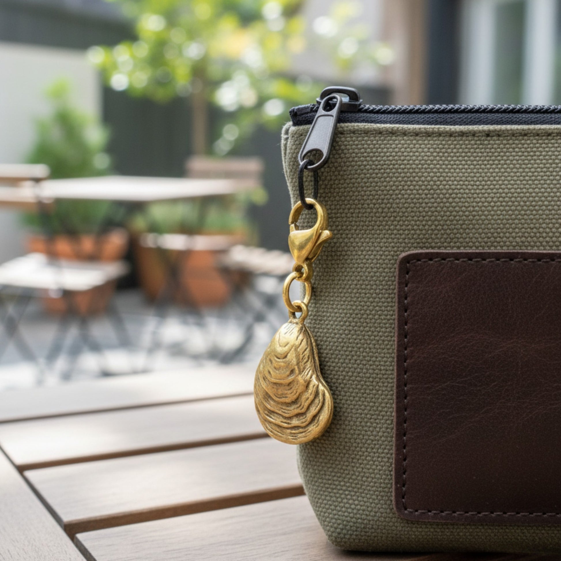 Hackberry Creek gold oyster shell clip-on charm attached to a green canvas zippered pouch on a wooden table.