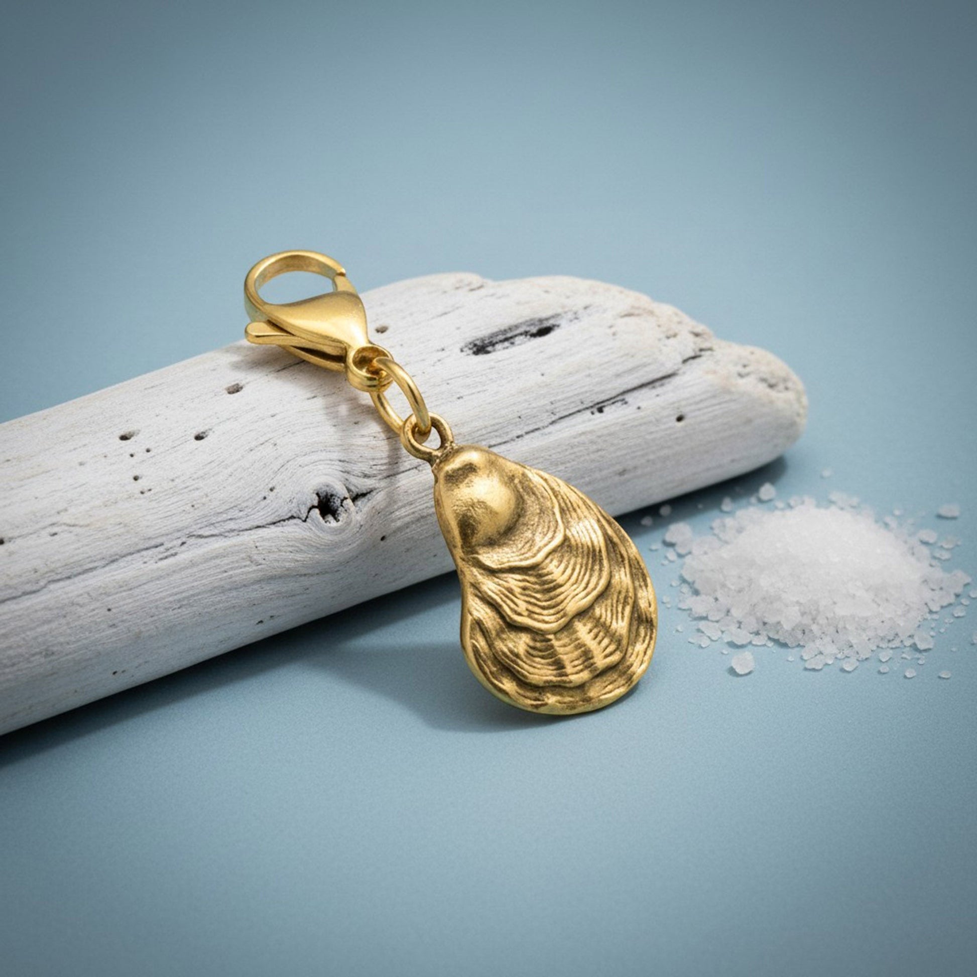 Hackberry Creek 24k gold-plated oyster shell charm with a lobster clasp, displayed on driftwood with sea salt.