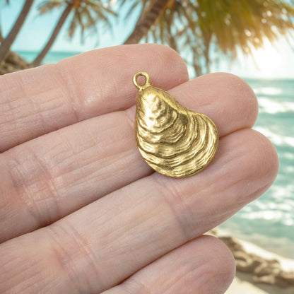 Hackberry Creek 24k gold plated oyster shell charm held in hand against a blurred tropical beach background