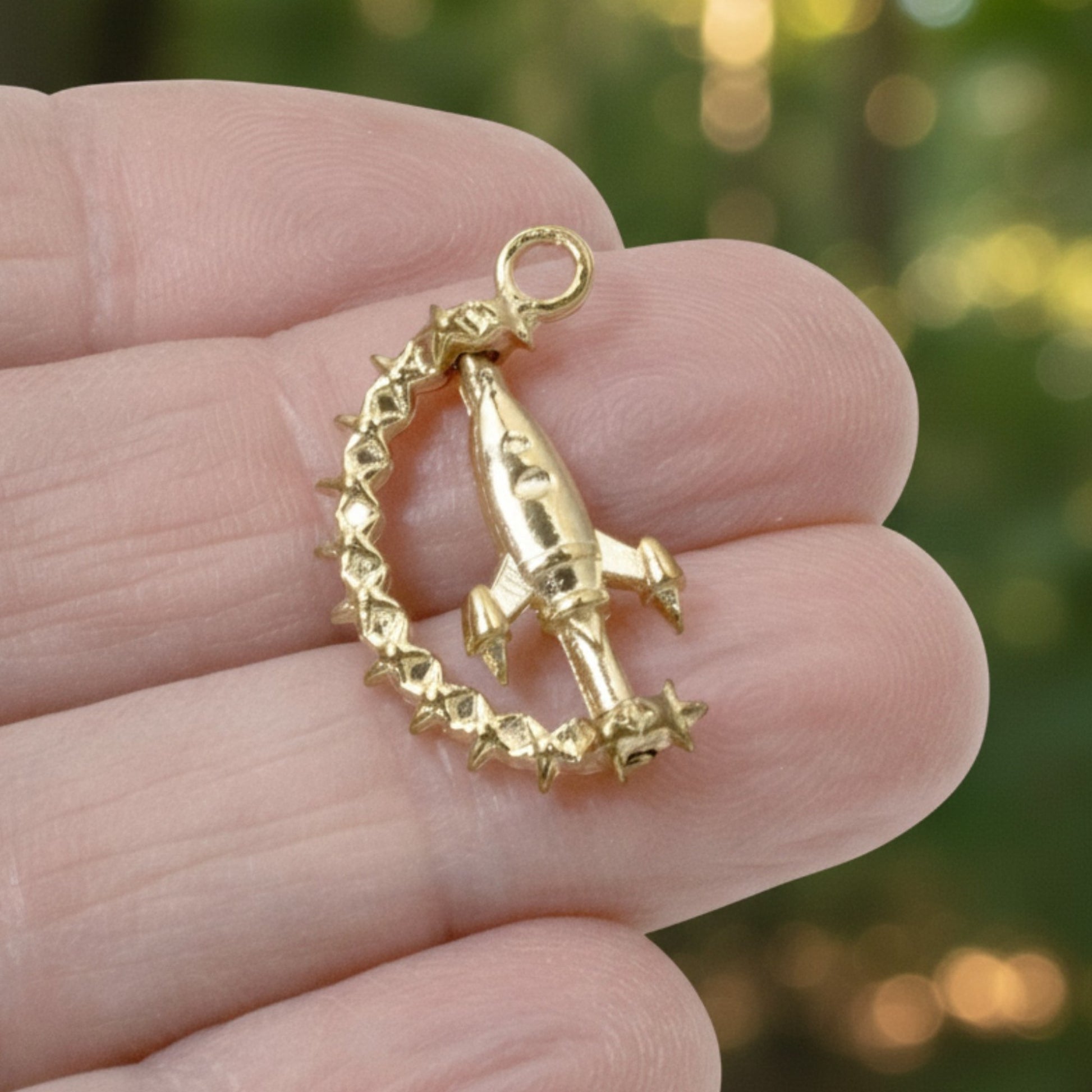 Hackberry Creek gold alloy spinning rocket charm with star details held in a hand against a blurred green background.