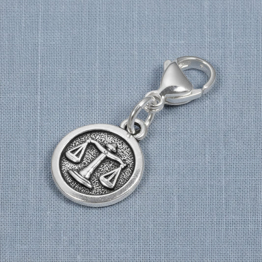 Hackberry Creek silver Libra zodiac charm with scales design and stainless steel lobster clasp on blue fabric.