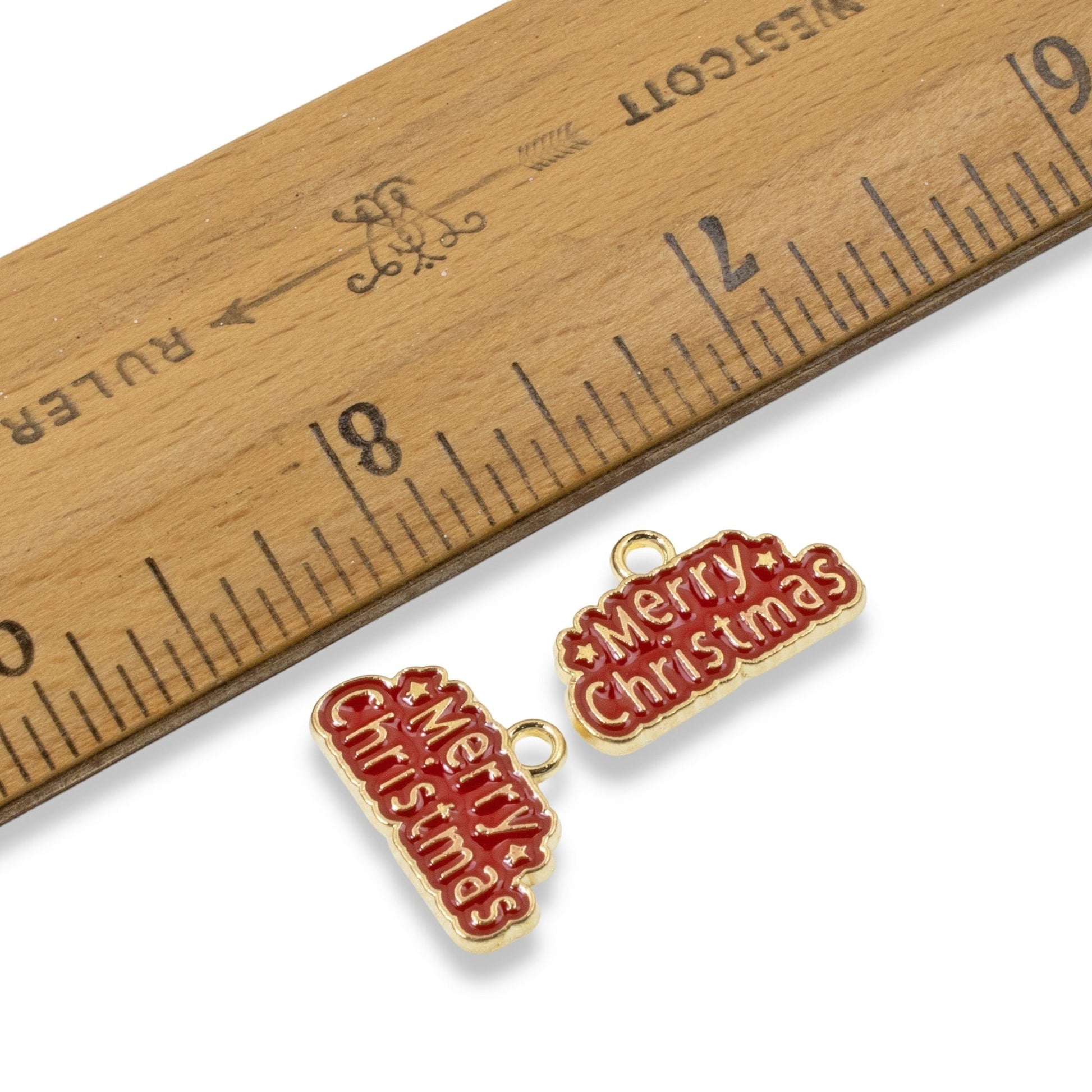 Hackberry Creek gold-plated metal alloy Merry Christmas charms with red enamel lettering next to a wooden ruler.