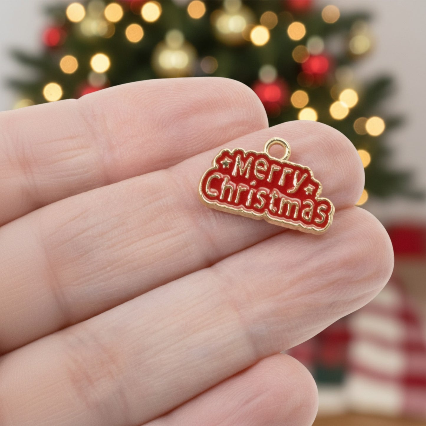 Hackberry Creek gold and red enamel Merry Christmas charm held in hand against a blurred festive tree background.