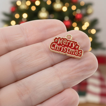 Hackberry Creek gold and red enamel Merry Christmas charm held in hand against a blurred festive tree background.