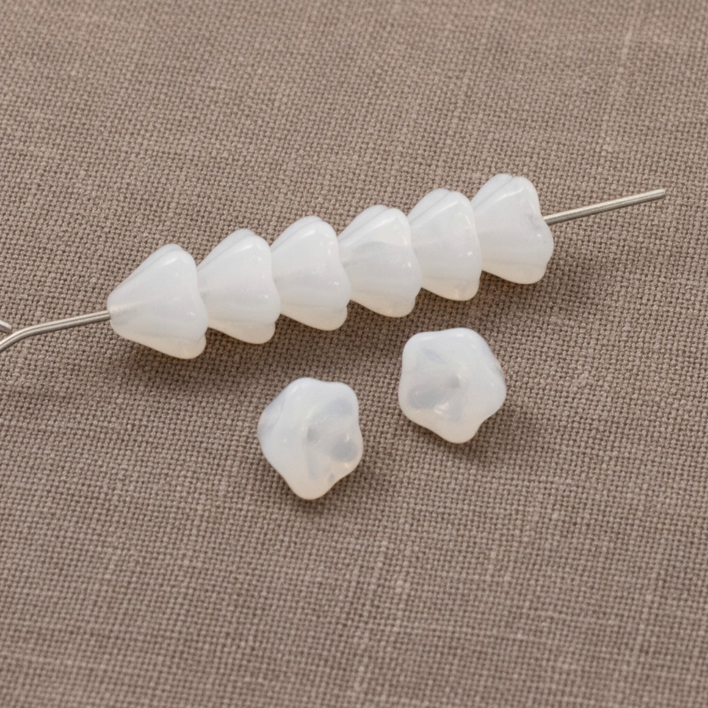 Hackberry Creek milky white Czech glass bell flower beads for jewelry making on a textured fabric background.