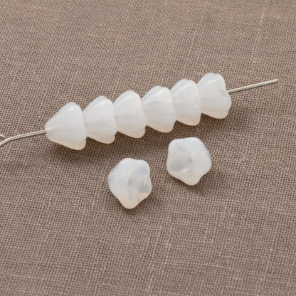 Hackberry Creek milky white Czech glass bell flower beads for jewelry making on a textured fabric background.