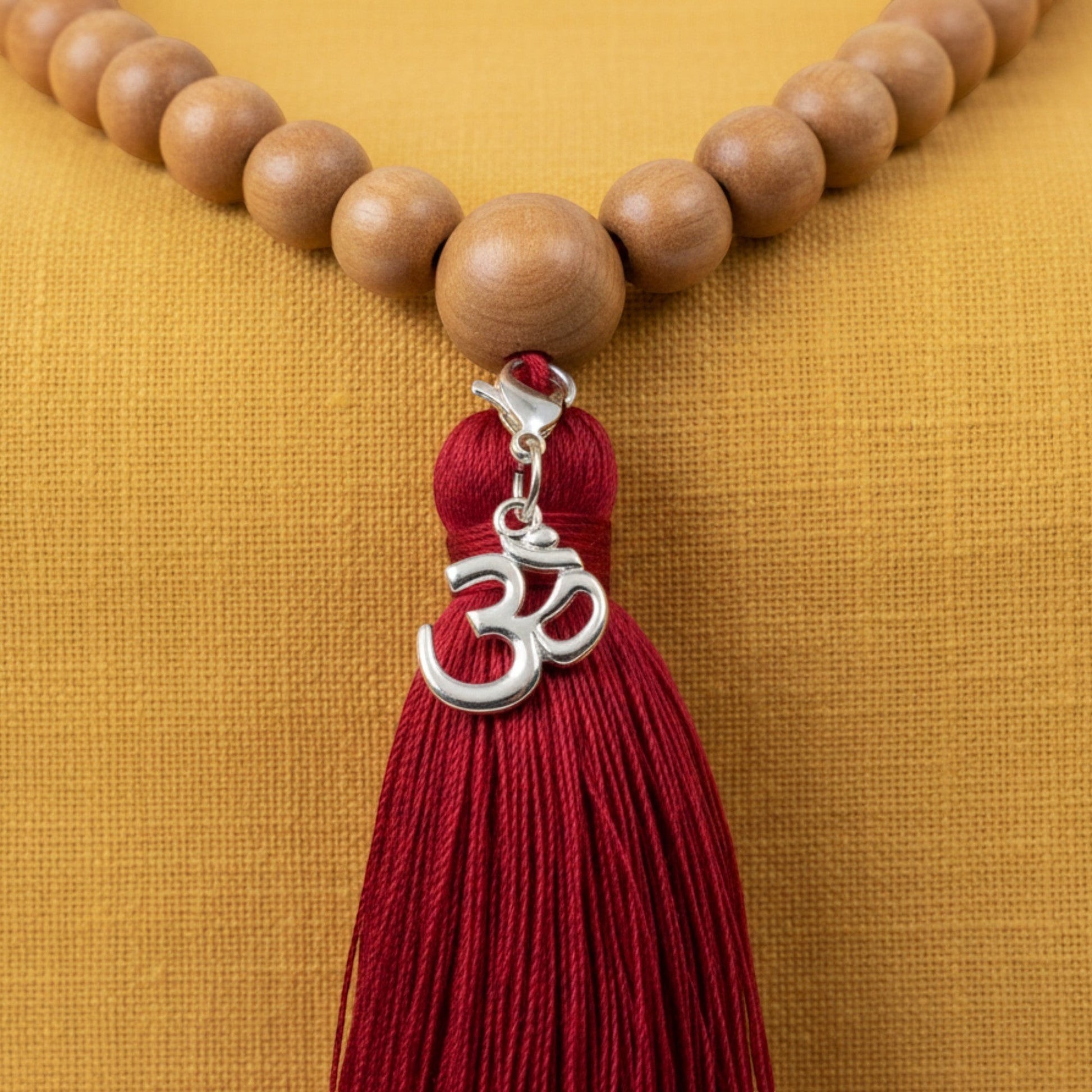 Hackberry Creek silver Om symbol charm with a deep red tassel hanging from wooden prayer beads on a yellow background