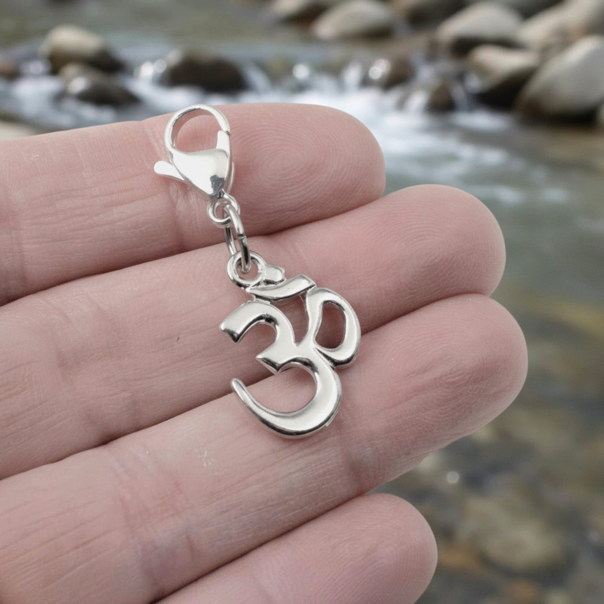 Hackberry Creek silver-toned Om symbol charm with a lobster clasp held in a hand against a blurred river background.