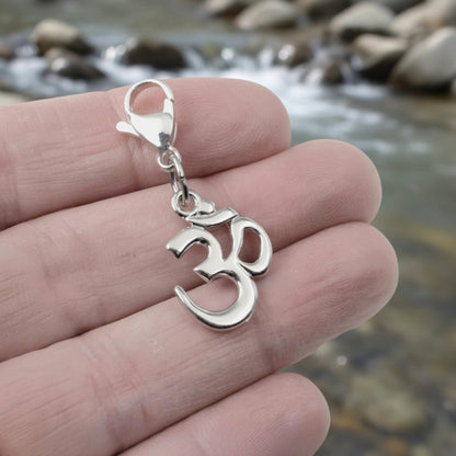 Hackberry Creek silver-toned Om symbol charm with a lobster clasp held in a hand against a blurred river background.