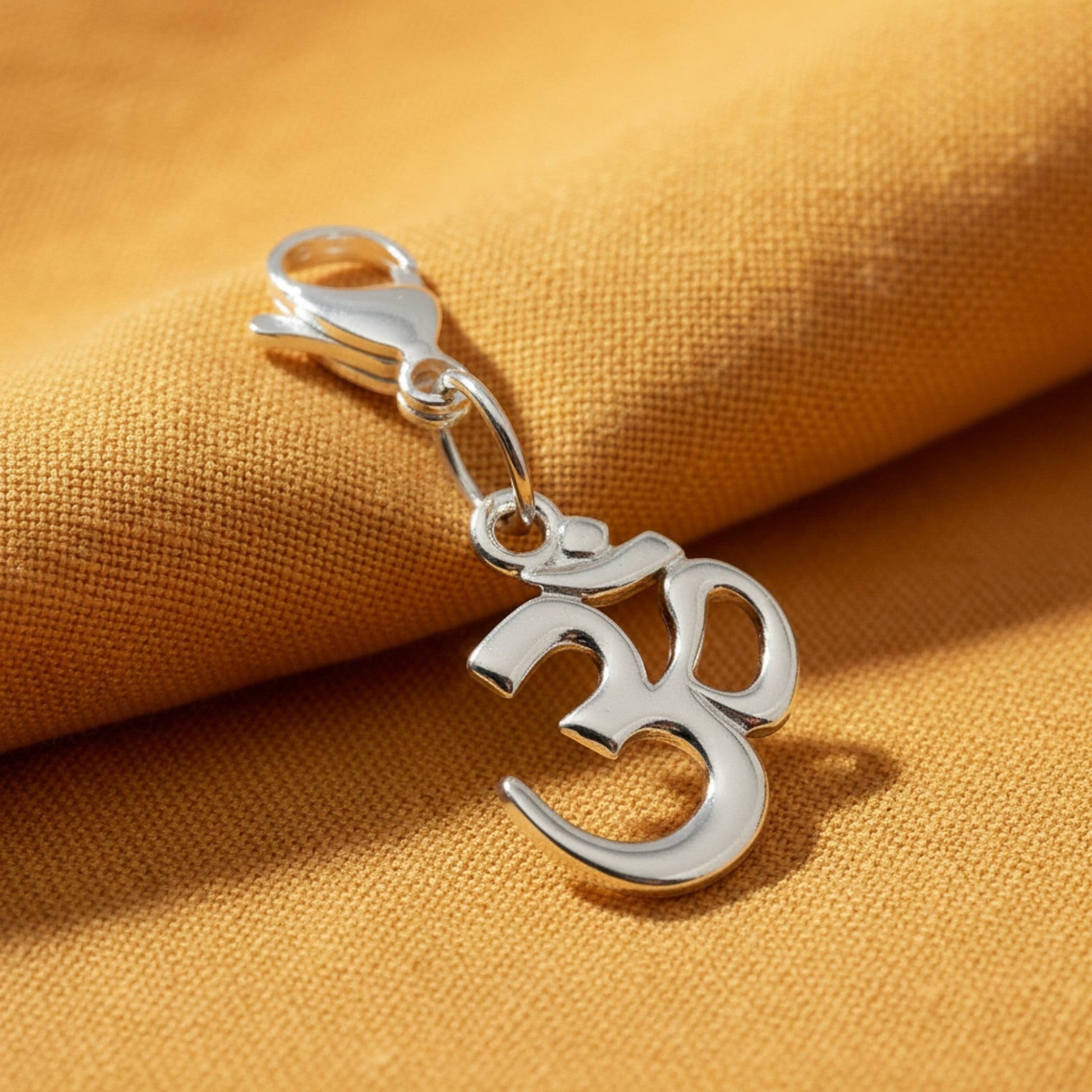 Hackberry Creek silver-toned pewter Om symbol charm with a lobster clasp, resting on a textured mustard yellow fabric.