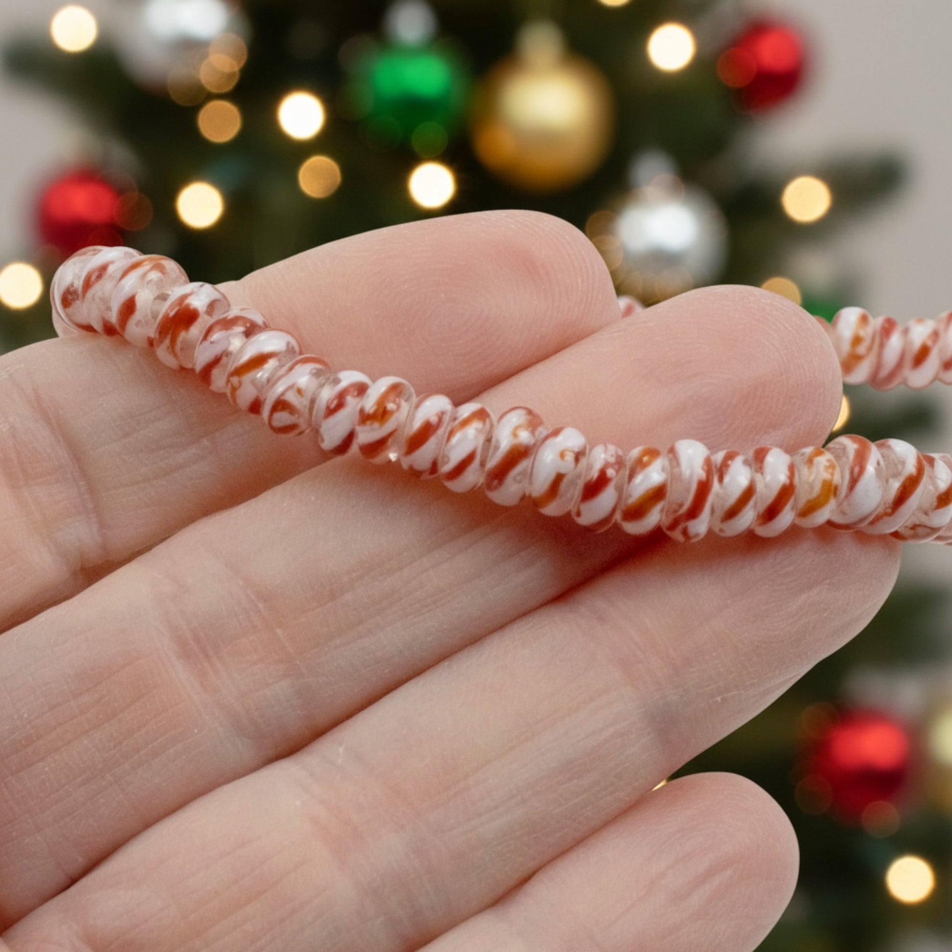 Hackberry Creek red and white peppermint stripe glass lampwork rondelle beads held in hand against a holiday tree