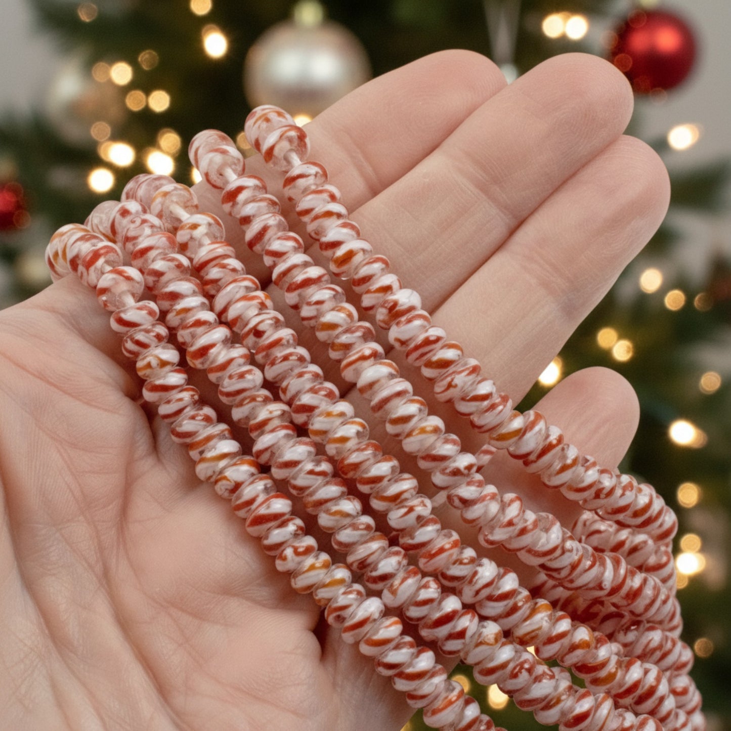 Hackberry Creek peppermint stripe glass rondelle beads on a strand held in hand against a blurred Christmas tree background
