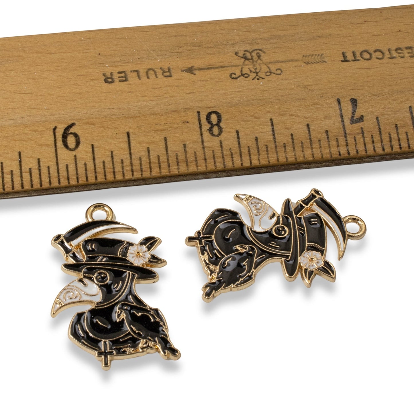 Hackberry Creek plague doctor raven head charms in black and white enamel with gold plating next to a wooden ruler.