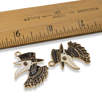 Hackberry Creek plague doctor raven head pendants in black and white enamel with gold plating next to a wooden ruler.