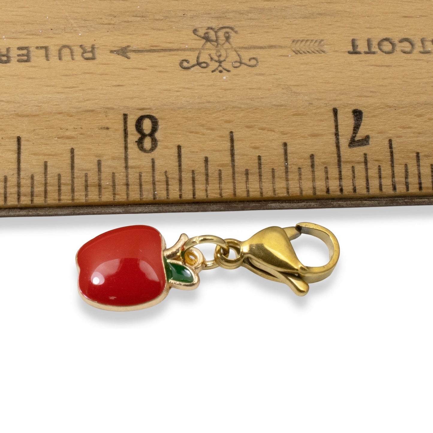 Hackberry Creek red apple charm with green leaf enamel and gold-plated brass clasp next to a wooden ruler.