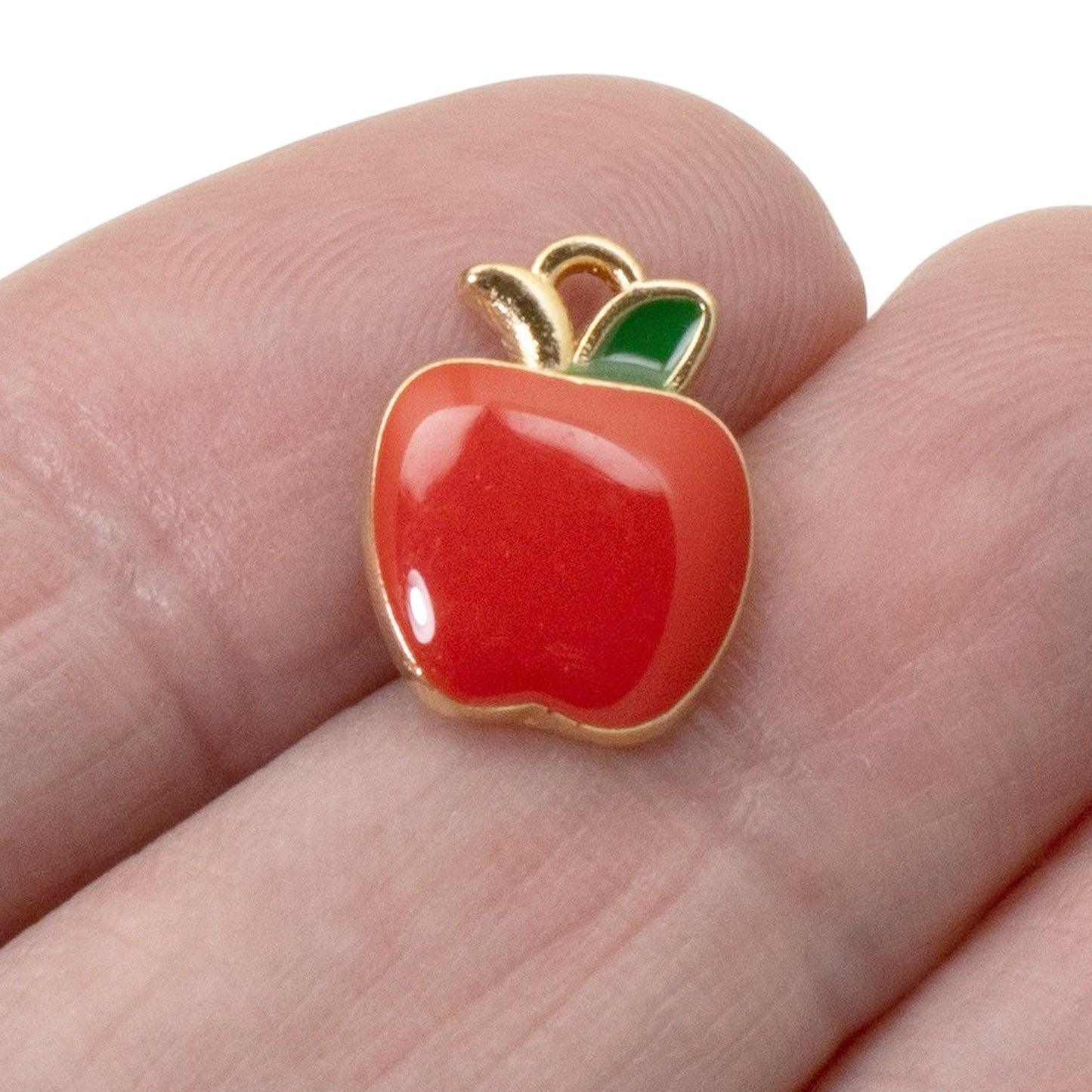 Hackberry Creek glossy red enamel apple charm with a green leaf and gold-plated metal edge held on a finger.