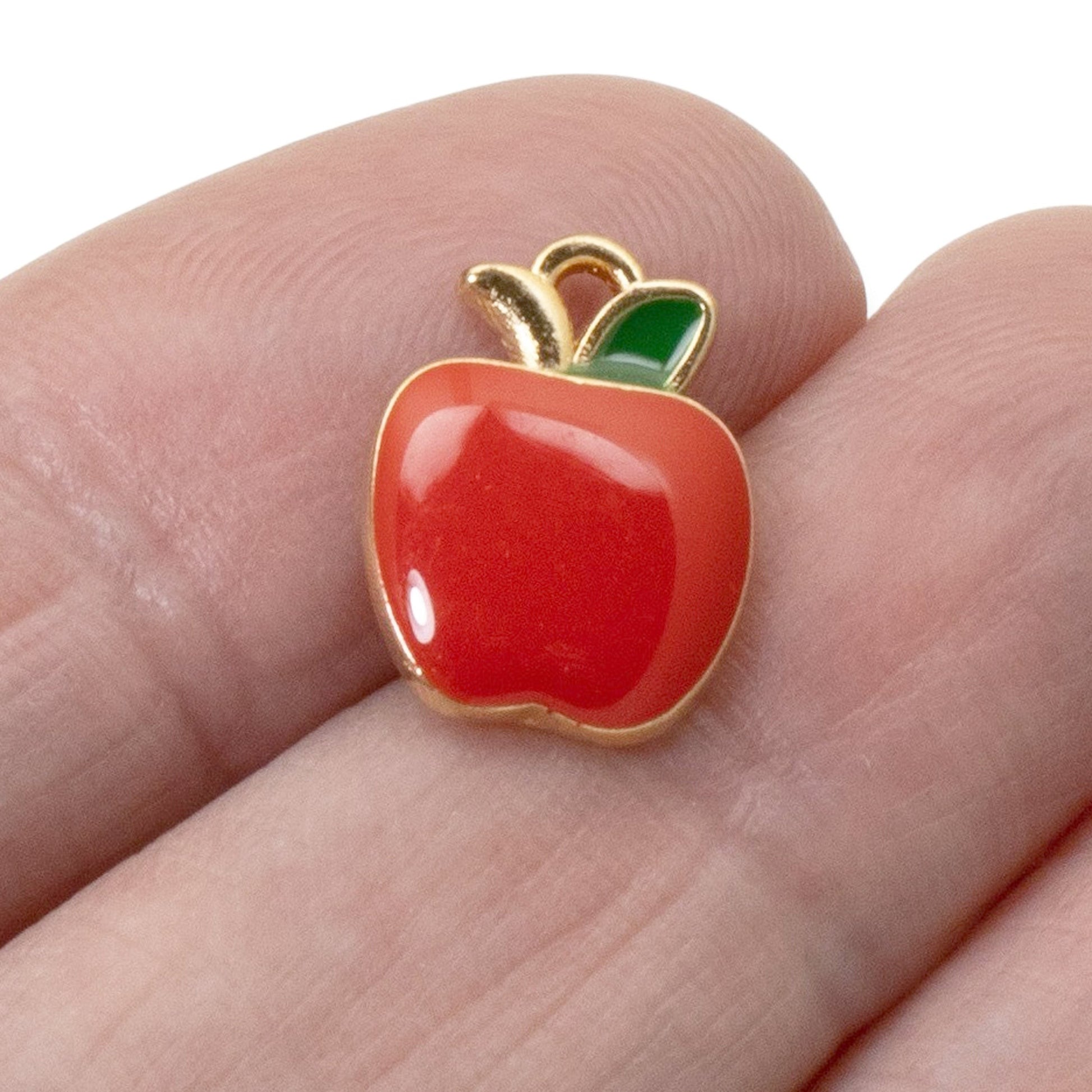 Hackberry Creek glossy red enamel apple charm with a green leaf and gold-plated metal edge held on a finger.
