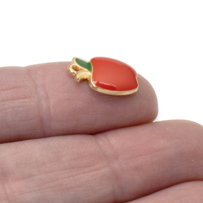 Hackberry Creek glossy red enamel apple charm with a green leaf, gold-plated metal alloy, held on a finger.