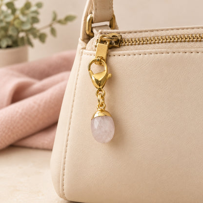 Hackberry Creek dainty oval rose quartz charm with a gold-plated stainless steel clasp attached to a beige handbag.