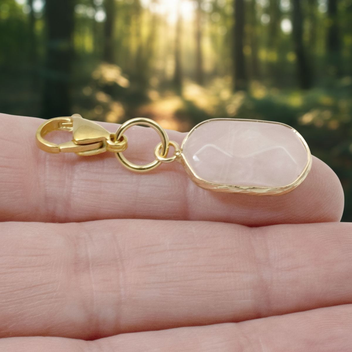Hackberry Creek oval rose quartz charm with gold-plated brass lobster clasp held in a hand against a forest background