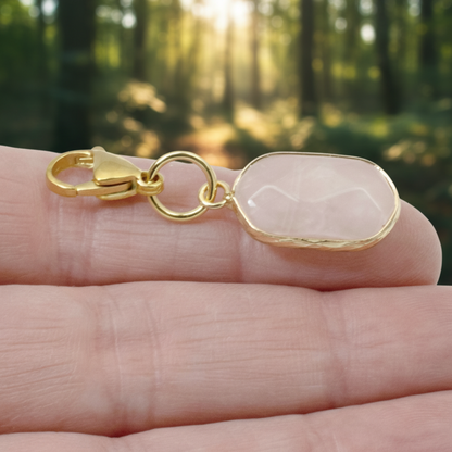 Hackberry Creek oval rose quartz charm with gold-plated brass lobster clasp held in a hand against a forest background