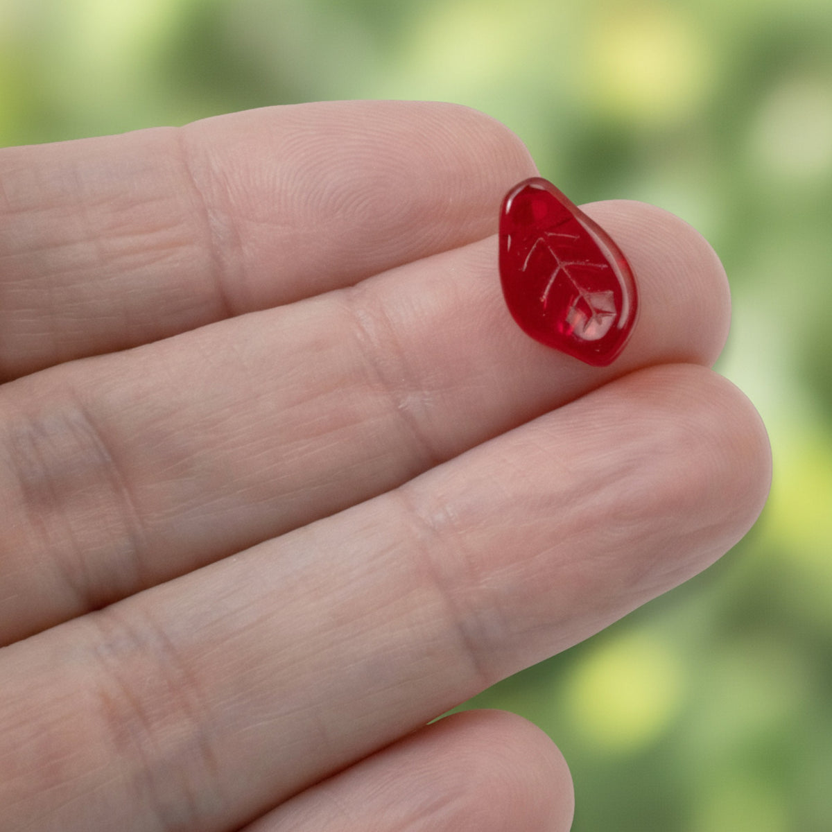 Hackberry Creek transparent Siam red Czech glass leaf bead with veining detail held on a fingertip