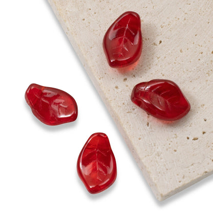 Hackberry Creek transparent Siam red Czech glass leaf beads with intricate veining on a textured stone surface.