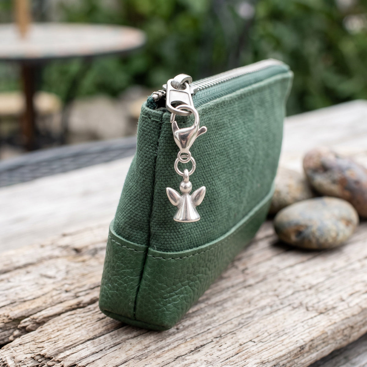 Hackberry Creek silver guardian angel charm clipped onto a green canvas and leather zipper pouch on a rustic table